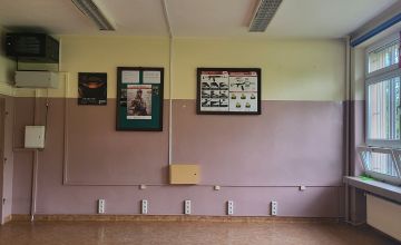 Pusta sala lekcyjna. Widok na ścianę w klolorze zgaszonego różu. Na ścianie wiszą dwie tablice z plakatami.